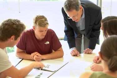 Eine Gruppe Studierende lernen gemeinsam an einem Tisch mit einem Ordner darauf. Ein Professor unterstützt die Studierenden beim Lernen.