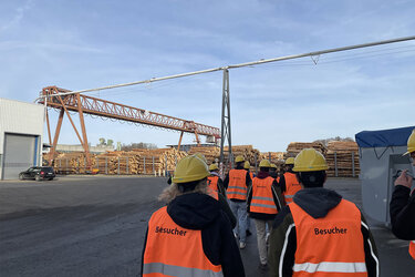 Studierende mit orangenen Warnwesten und gelben Helmen laufen über das Werksgelände