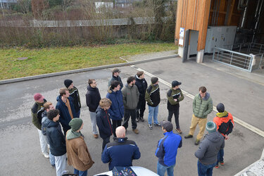 Studierende stehen in einem Kreis und hören dem Referenten zu