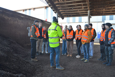 Studierende stehen im Kreis und hören dem Referenten zu.