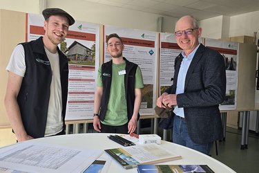 Ein Professor, ein Studiengangskoordinator und ein Student stehen an einem Infostand und lächeln in die Kamera.