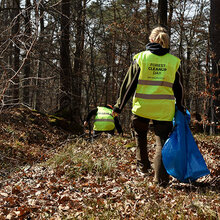Eine Frau läuft mit einem blauen Müllsack durch den Wald und sammelt Müll ein