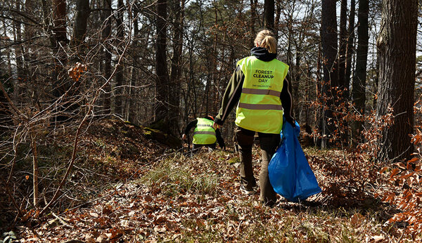 Eine Frau läuft mit einem blauen Müllsack durch den Wald und sammelt Müll ein