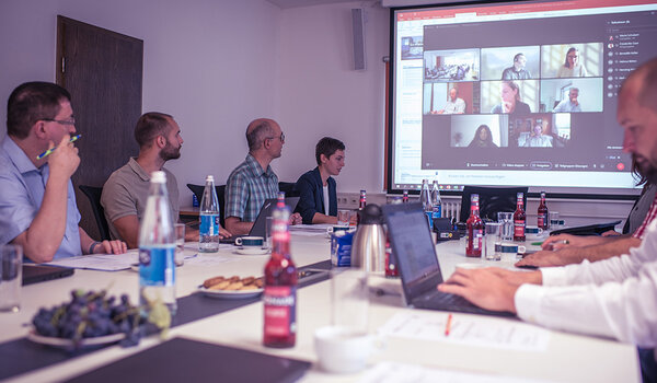 Teilnehmer der Sitzung sitzen an einem Konferenztisch. Die Onlinteilnehmer der Sitzung sind auf die Leinwand projeziert.