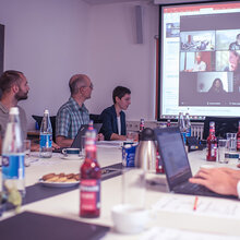 Teilnehmer der Sitzung sitzen an einem Konferenztisch. Die Onlinteilnehmer der Sitzung sind auf die Leinwand projeziert.