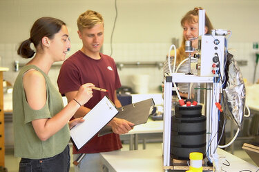 Zwei Studentinnen und ein Student betrachten gemeinsam einen Versuch im Labor und machen sich Notizen.
