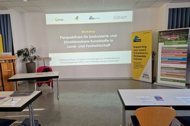 Blick in den noch leeren Veranstaltungsraum. Auf der Leinwand ist die Begrüßungsfolie zu sehen