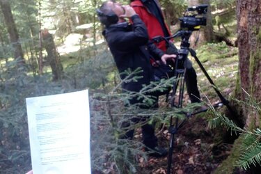 Kameramann und Referent stehen an einem Baum und schauen in die Krone. Im Vordergrund hält eine Hand das Script in das Bild.