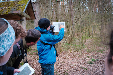 Der Professor zeigt auf einem ausgedruckten Luftbild den Studierenden im Wald den Standort