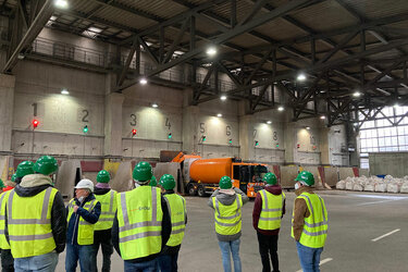 Studierende mit grünen Helmen und neongelben Warnwesten stehen in einer großen Werkshalle