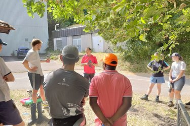 Studierende stehen in einem Kreis und hören dem Referenten zu