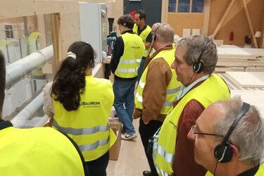 Teilnehmer stehen mit gelben Warnwesten in einem Sägewerk. Mit Kopfhörern hören sie dem Referenten zu