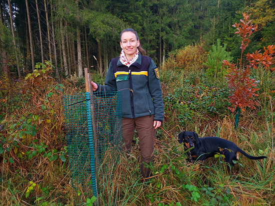 Frau Verena Strasdeit steht mit ihrem Hund im Wald und lächelt in die Kamera